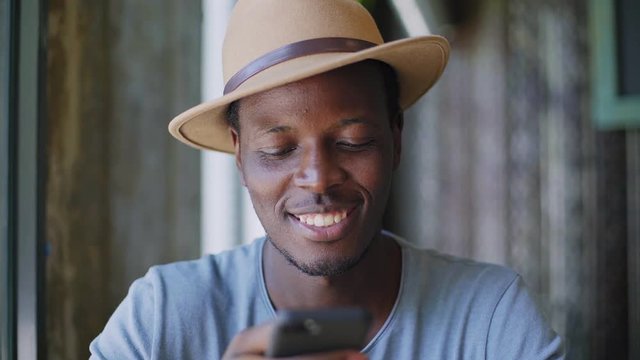 Portrait shot of smiling and laughing young black man texting message or email in his smartphone, sitting in cafeteria at lunch time and wearing trendy fedora hat at summer sunny day