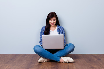 Young pretty smiling woman in checkered shirt and jeans sitting on the floor and studying using her laptop