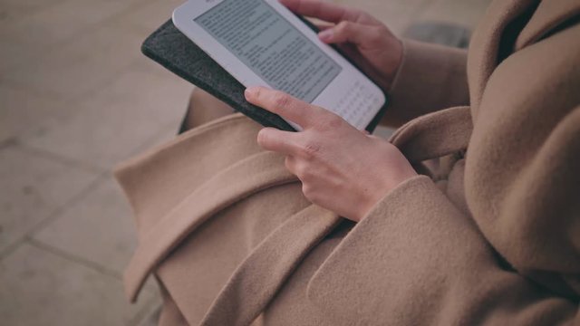 zoom out shot of trendy brunette girl in fashionable winter outfit in park, dreaming while reading her white technologic reading device instead of paper book and dives into world of future technology