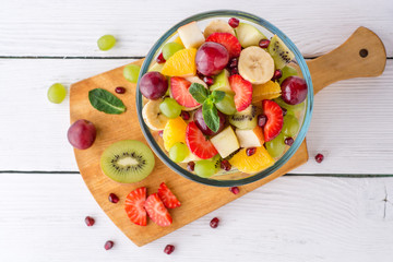 Healthy fresh fruit salad in glass bowl on white wooden background.