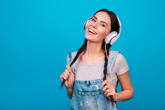 Portrait of cute young girl with funny pigtails in jeans overalls listening to music against blue background