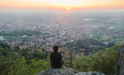 Man sitting on a rock on a hill above the city meditating and watching the sunset - horizontal