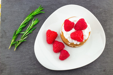 Fresh Red Raspberries on a white artistic plate / Red Fresh Raspberries on a white artistic plate with cookies, rosemary, chocolate and cream in the background.