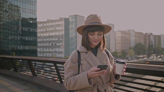 Dreamy beautiful young woman chats with her friends using messaging apps on her futuristic smartphone surround by busy urban city environment