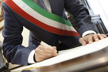 foto b/n della mano di un sindaco con fascia tricolore che firma con una penna un documento