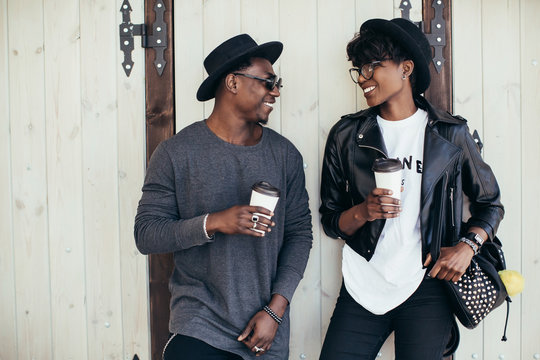 Stylish and young african couple drinking coffee and speaking near a wooden wall.