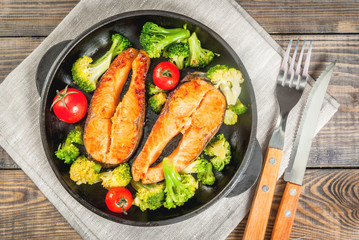 Healthy eating, diet. Baked grilled trout (salmon) with vegetable garnish - broccoli, tomatoes. In a portioned frying pan, on a wooden table. Top view copy space