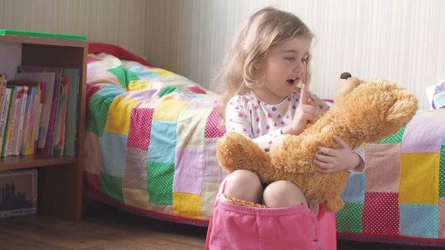 Little girl sitting on the potty beside the bed and plays with a Teddy bear.