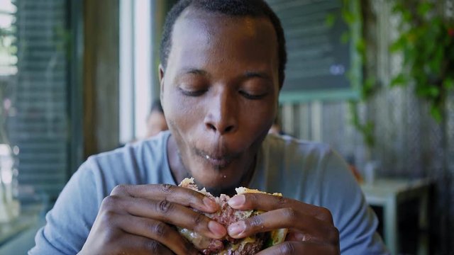 Hand held shot of a stylish black american man devouring and biting on a chewy enormous hamburger with two steaks, he smiles and grins into the camera full of joy and satisfaction