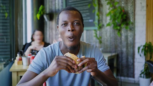 A hand held shot of a young hipster and contemporary black teenager filling himself with typical american fast food, a cheeseburger he munches on with hunger and appetite