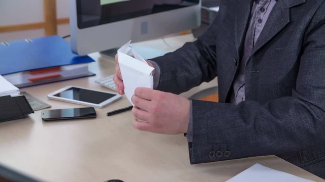 A businessman is taking some money out of the envelope, then he is putting it back inside. Afterwards, he is closing the envelope and putting it into the pocket of his jacket.
