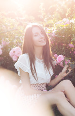 Obraz na płótnie Canvas Красивая девушка с длинными волосами сидит на траве в парке возле розовых роз, теплая тонировка 