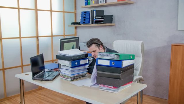 A young businessman is sitting behind his desk in the office and he is overwhelmed with work. He throws papers up in the air and watched them falling down on the floor.
