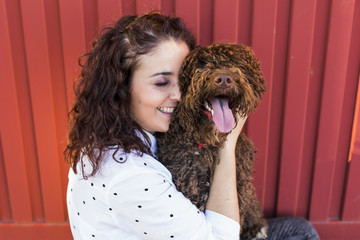 Beautiful young woman hugging her dog, a brown Spanish water dog over red background. She is...