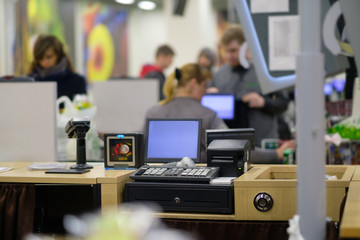 Empty cash desk with computer screen and card payment terminal with cashiers serves customers on background