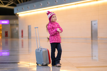Adorable girl in winter outfit inside of airport building with roller suitcase posing