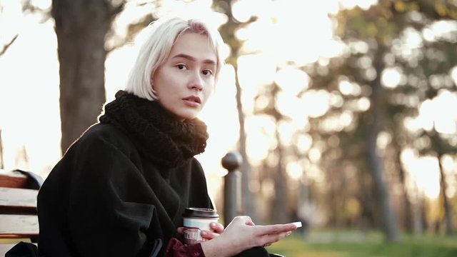 Blonde woman girl outside. In the hands of a smartphone and coffee. Sits on a wooden bench. He looks at the smartphone, turns abruptly at the camera. Surprise on the face. Called