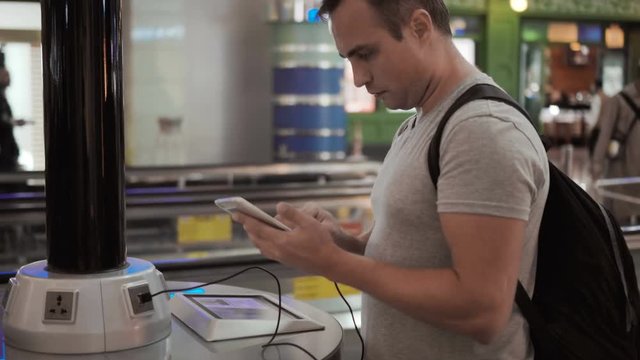 Handsome young man in airport terminal. Standing near charging stand pluging in smartphone