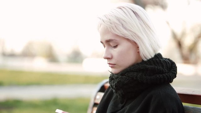 Blonde woman girl outside. Looks at the smartphone. Sits on a wooden bench. Dials text in the smartphone, a serious facial expression. Emotionless. Side view. Pump