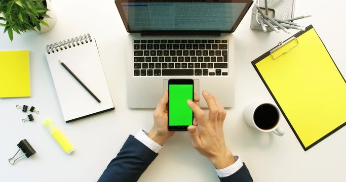 Business man using smart phone with green screen, scrolling pages, tapping on touch screen on office desk background. Hands top view. Green screen. Chroma key