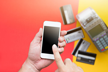 Human hand holding Smartphone for payment with blurred credit card and credit card machine on red and yellow background.