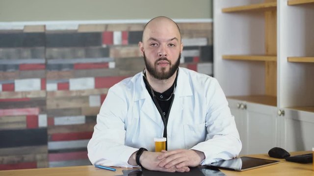 a portrait of a adult 30s bald bearded doctor sitting on his medical room office among table 4k