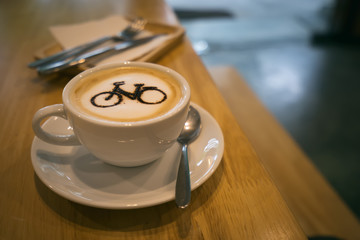 A cup of cappuccino with intricate bicycle latte art on a wooden table captures the blend of creativity and coffee culture in a cozy cafe setting.