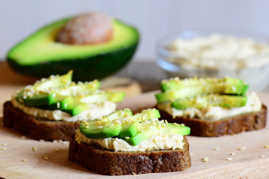 Homemade chickpeas hummus and avocado sandwiches on a wooden board, avocado half, hummus in a glass bowl. Vegetarian open sandwiches made with rye bread, avocado slices, hummus and fried sesame seeds