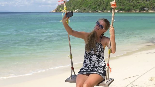 Smiling girl taking photo with camera phone on a swing at beach.