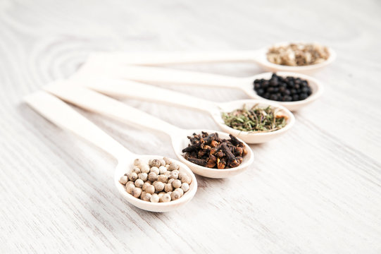 Various spices and herbs in wooden spoons on white table background. Aromatic food cooking ingredients. Black and white pepper, clove, savory, fennel seeds. Soft focus.