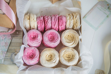Нежные кремовые и розовые зефир, упакованные в коробочки из крафтовой бумаги с атласными лентами на белом деревянном столе.