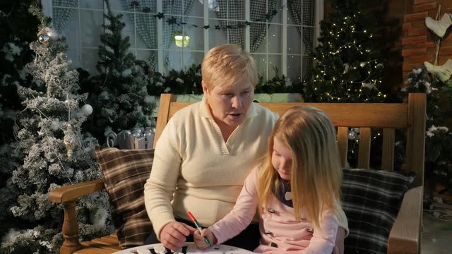 Grandma and Little Girl Drawing Postcards Together in a Studio Brightly Decorated With Lights Garlands Firs and Traditional Attributes of Christmas