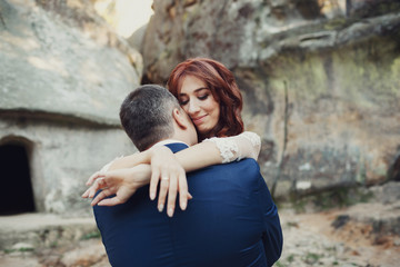 Groom in blue suit hugs bride tender standing before grey rocks