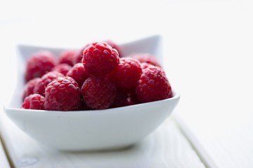 Fresh and slightly imperfect raspberries in a white bowl on white board table