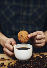 Close-up of male hands holding coffee mug and cookie on table with coffee beans. 