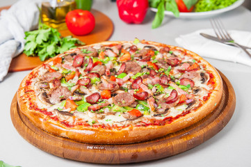 Selective focus close up Delicious Meat Pizza with bacon and sausages on the wooden board on the kitchen table with different bright ingredients