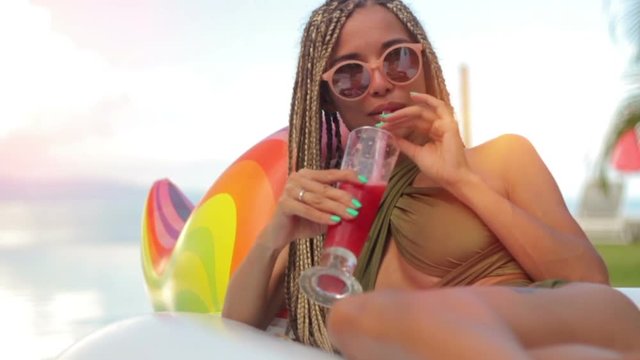 Attractive woman drinking red cocktail and sitting on Inflatable unicorn in swimming pool on sea background