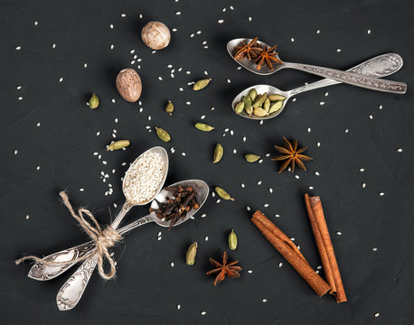 Various spices in metal vintage spoons and cinnamon sticks on dark rustic background. Cardamom, nutmeg, anise, clove, sesame seeds. Aromatic food cooking and baking ingredients.