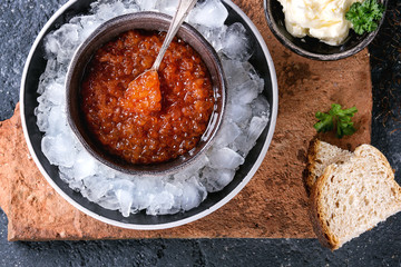 Bowl of red caviar on ice with spoon served with sliced bread, butter and herbs on terracotta board over black stone texture background. Top view, close up