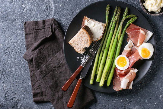 Cooked green asparagus with half boiled egg, sliced bread, butter and ham bacon served with sea salt and cutlery on black ceramic plate over dark stone texture background. Top view, fine dining