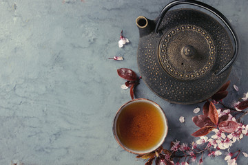 Obraz na płótnie Canvas Black iron teapot and traditional ceramic cup of tea with blossom pink flowers cherry branch over gray blue metal texture background. Top view with space, Asian style.