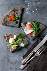 Black pancakes crepes with variety of salt filling red caviar, ham, fish salmon pate, green salad with vintage cutlery over gray texture metal background. Top view