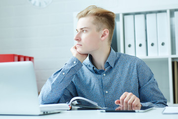 Handsome pensive young business man sitting at the dask with laptop and looking at the window. © cameravit