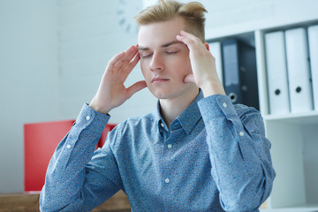 Stressed and tired. Young caucasian man massaging temple and keeping eyes closed while sitting at his working place.