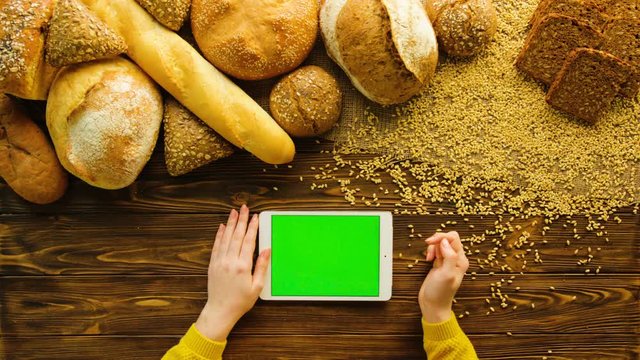 Woman using tablet device with green screen on wooden table with fresh bread. Bakery background. Top view. Female hands scrolling pages, tapping on touch screen. Close up. Chroma key