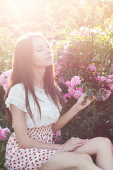 Obraz na płótnie Canvas Красивая девушка с длинными темными волосами сидит на траве в саду возле куста розовых роз