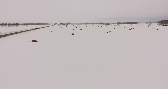 Siberia, 2017: Aerial view from helicopter: countryside in winter. A secluded road, a snowy field with haystacks. Taken from the air: movement along the winter field. Sleeping winter nature