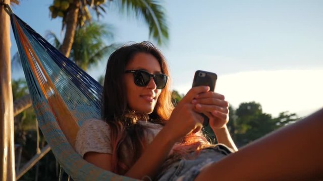 Atractive Young Hipster Girl Using Mobile Phone in Hammock at the Beach near the Sea at Sunset. Koh Phangan, Thailand. HD Slowmotion.