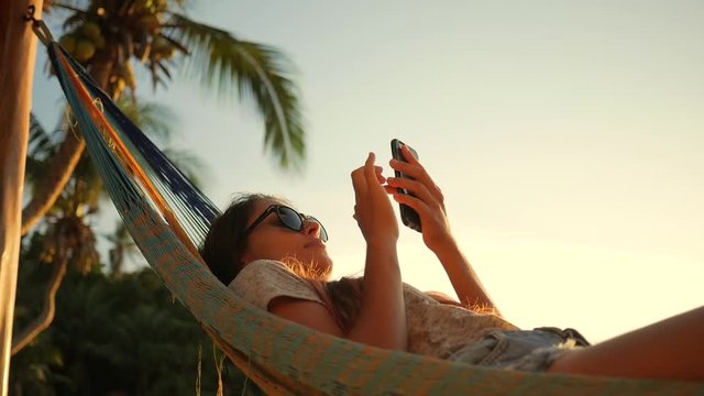 Relaxed Mixed Race Young Woman Looking at Mobile Phone in Hammock at the Beach near the Sea at Sunset. Koh Phangan, Thailand. HD Slowmotion.