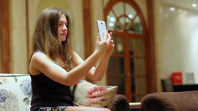 A nice brunette does selfie on her new smartphone in the lobby of an expensive hotel. Beautiful girl is making a photo while sitting on the couch.